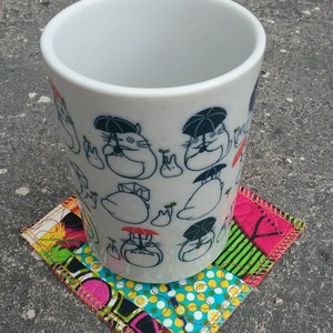 May include: White ceramic mug with a black and white illustration of cartoon characters holding umbrellas. The mug is sitting on a colorful patchwork fabric coaster.