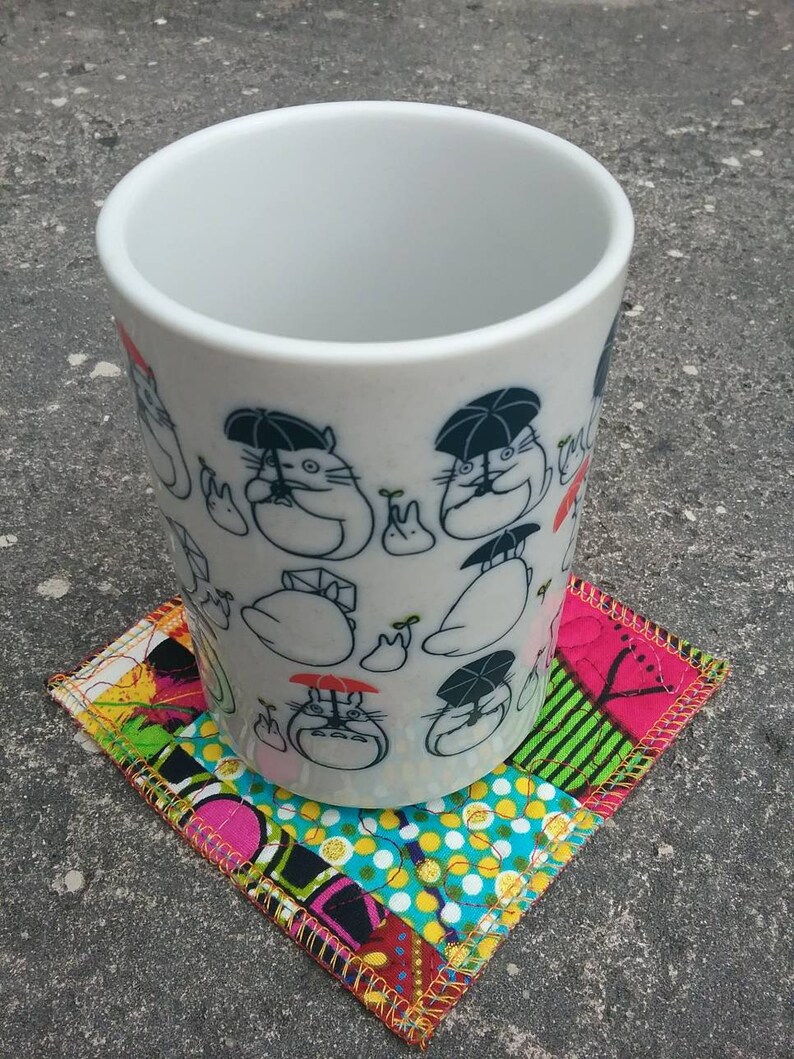 May include: White ceramic mug with a black and white illustration of cartoon characters holding umbrellas. The mug is sitting on a colorful patchwork fabric coaster.