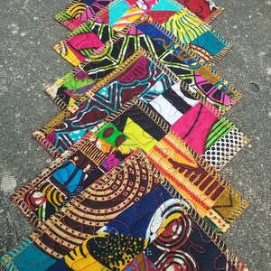 May include: A set of colorful fabric coasters with a patchwork design. Each coaster is made of different fabrics with geometric patterns in shades of yellow, blue, red, green, and black. The coasters are edged with gold trim and have a quilted texture.