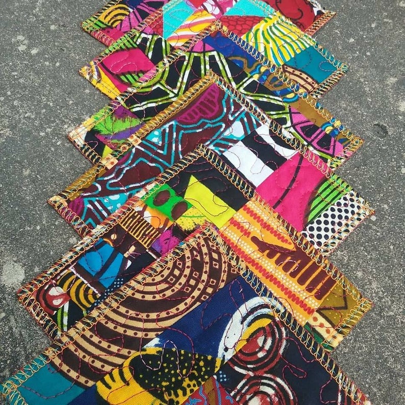 May include: A set of colorful fabric coasters with a patchwork design. Each coaster is made of different fabrics with geometric patterns in shades of yellow, blue, red, green, and black. The coasters are edged with gold trim and have a quilted texture.