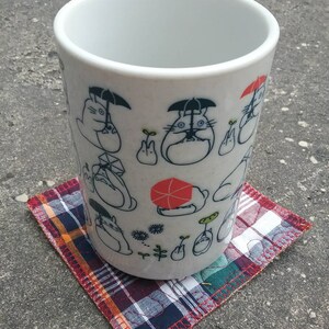 May include: White ceramic mug with a black and white cartoon design featuring a character with an umbrella. The mug is sitting on a red, blue, and white plaid coaster.