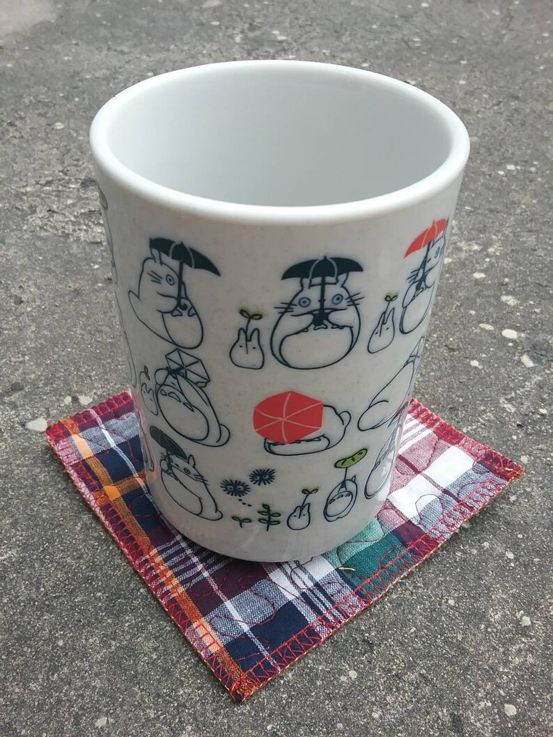 May include: White ceramic mug with a black and white cartoon design featuring a character with an umbrella. The mug is sitting on a red, blue, and white plaid coaster.