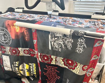 May include: A close-up of a quilt being quilted on a longarm quilting machine. The quilt is made of various fabrics, including black and red patterned fabric, and features several patches with text, including "We Gotta Know", "World Peace", "Show You No Mercy", "Malfunction", "Street Justice", "Survival Of The Fittest", "Don't Work The Limit", "Live The Truth", "And The Rest", "Don't Fade Me", "Out Of The Own", "Some Of The Times", "PROTK", "VA MERU", "Flags", and "The Age Of Qu".