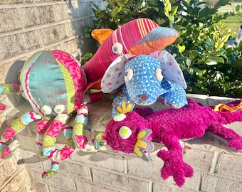 May include: Four colorful plush toys, including a pink and orange dolphin, a blue and white dog, a pink and green octopus, and a pink and yellow lizard. The toys are made of fabric and have large, expressive eyes.