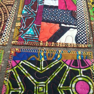 May include: Set of four colorful fabric coasters with a patchwork design. Each coaster features a different pattern of bright colors and geometric shapes. The coasters are edged with a gold trim.