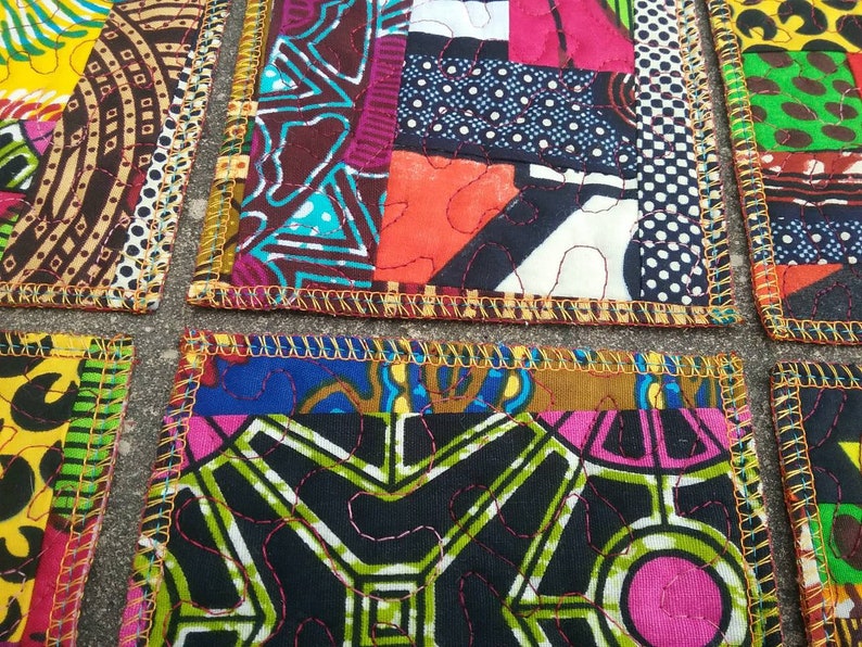 May include: Set of four colorful fabric coasters with a patchwork design. Each coaster features a different pattern of bright colors and geometric shapes. The coasters are edged with a gold trim.