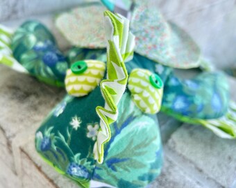 May include: A green and blue fabric toy with a green ruffled horn, green eyes, and a floral pattern. The toy is sitting on a white surface.