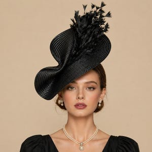 May include: A black, woven hat with an unusual, asymmetrical shape and feather detailing. The hat is worn by a person wearing a black top and pearl jewellery. The background is a plain, neutral colour.