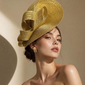 May include: A metallic gold fascinator hat featuring a wide, circular brim and a prominent, sculptural bow detail on the side. The hat is constructed from a textured, woven material with a shimmering finish, positioned at an angle on the head.
