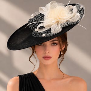 May include: A wide-brimmed black hat featuring a decorative arrangement of cream-coloured fabric flowers, sheer netting, and small white faux pearls. The hat has a structured, flat brim and a rounded crown, creating a bold and formal headpiece.