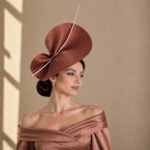 May include: A terracotta-coloured, wide-brimmed hat with an unusual, asymmetrical shape. The hat has a prominent bow detail and a single, long white feather. This hat is a stylish accessory, suitable for special events.