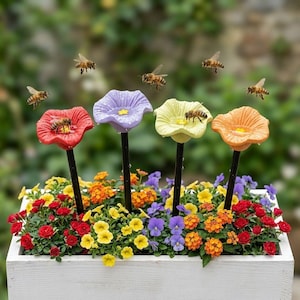 Può includere: Quattro picchetti decorativi da giardino a forma di fiori rossi, viola, gialli e arancioni sono inseriti in una fioriera in legno bianco riempita di piccoli fiori rossi, gialli, arancioni e viola, con diverse api che volano attorno ai fiori.