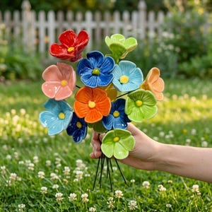 Puede incluir: Un ramo de flores de cerámica que presenta una variedad de colores, incluyendo rojo, naranja, azul, azul claro, verde y melocotón. Cada flor tiene cinco pétalos redondeados y un pequeño centro amarillo, unidos a tallos oscuros.