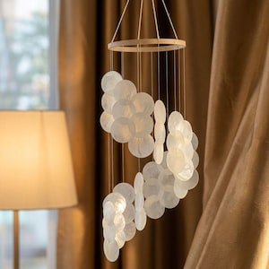 May include: A decorative wind chime featuring numerous white, round, and oval shell pieces suspended from a wooden ring. The shells are strung on thin cords, creating a cascading effect. The background includes a lamp and curtains.