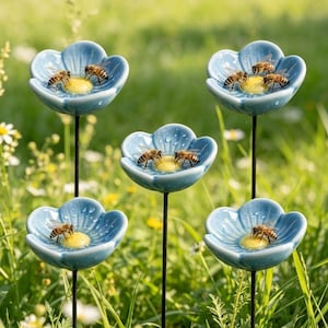 Könnte beinhalten: Fünf blaue, blumenförmige Bienen-Wasserfütterer aus Keramik mit schwarzen Stielen werden in einer grasbewachsenen Umgebung im Freien gezeigt. Jede Blume enthält eine gelbe Mitte und mehrere Bienen. Die Blumen haben eine hellblaue Glasur.