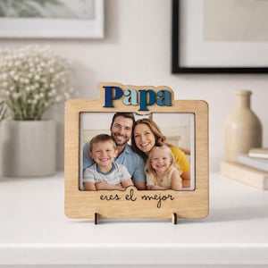 Puede incluir: Marco de fotos de madera con la palabra "Papa" en letras azules y verdes en la parte superior. El marco contiene una foto familiar y las palabras "eres el mejor" están escritas en la parte inferior. El marco se muestra sobre una superficie blanca.