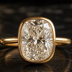 May include: A gold ring featuring a large, rectangular-cushion cut diamond. The diamond is set in a bezel setting, and the band is a simple, polished gold. The ring is displayed on a dark, textured surface.