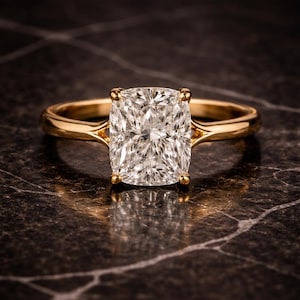 Puede incluir: Un anillo de oro con un gran diamante de talla cuadrada. El diamante claro y brillante está engastado en una montura de cuatro garras. La banda de oro pulido contrasta con la superficie oscura y veteada.