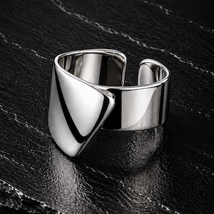 May include: A polished silver-tone ring featuring a wide, open-ended band with a prominent, smooth, triangular-shaped overlapping front section. The metal surface is highly reflective, creating sharp highlights and deep shadows across its curved form.