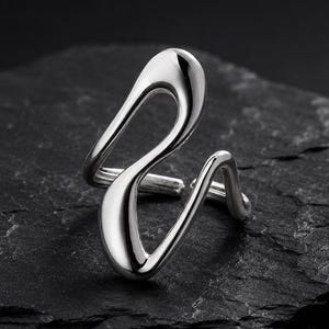 May include: A polished silver-toned ring featuring an open, fluid, and asymmetrical design with smooth, rounded edges. The band consists of a continuous, curving metal form that creates a hollow, abstract shape, showcasing a reflective surface.