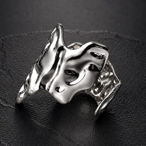 Peut inclure: Une bague argentée présentant une surface irrégulière rappelant du métal fondu avec des contours organiques et fluides. Le métal possède une finition hautement polie et réfléchissante qui souligne le design texturé et inégal de l'anneau ouvert.