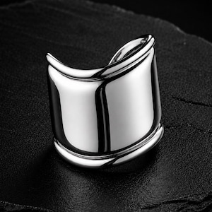 May include: A wide, polished silver-tone ring with a smooth, curved surface and raised edges along the top and bottom. The metal has a high-shine, reflective finish that catches the light, creating distinct highlights across its rounded, open-ended band.