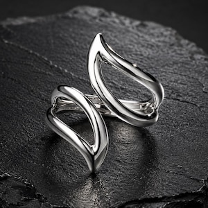 May include: A polished silver-tone ring featuring an open, bypass-style design with two distinct, teardrop-shaped loops that curve towards each other. The metal surface is smooth and reflective, creating a continuous, fluid silhouette.