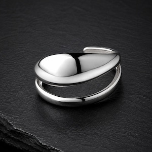 May include: A polished silver ring featuring an open, bypass-style design. The ring consists of two distinct bands that curve and meet at a smooth, rounded, teardrop-shaped focal point, creating a layered and reflective metallic appearance.