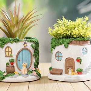 Puede incluir: Dos macetas decorativas de cerámica blanca con forma de casa, adornadas con hiedra verde y puertas y ventanas en miniatura. Una presenta una puerta azul y una estatua de perro, la otra tiene una puerta marrón y una estatua de perro. Cada maceta contiene una planta diferente.