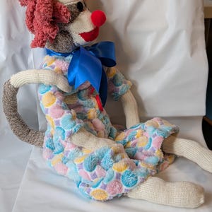 May include: A handmade clown doll with a red nose, red curly hair, and a blue satin bow. The doll wears a colorful plush outfit with pastel polka dots. The arms and legs are made of a cream-colored knit fabric.