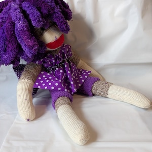 May include: A handmade sock doll with a purple yarn wig, a red mouth, and a purple polka dot dress. The doll has cream-colored feet and arms, and brown and cream-colored legs and torso. The doll is lying on a white surface.
