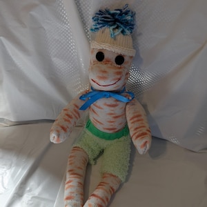 May include: A handmade sock monkey doll with a cream-colored knit hat and a blue pom-pom. The doll has black button eyes, a stitched smile, and a blue ribbon bow. It wears green shorts and orange and white striped socks.