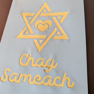 May include: A light blue rectangular stencil featuring a yellow Star of David with a small heart in the center. Below the star, the text "Chag Sameach" is cut out in a cursive font. The stencil is made of a flexible material with visible cutouts.