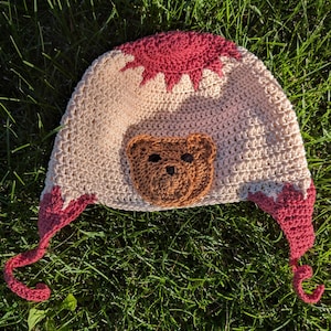 May include: A handmade crochet hat in cream and burgundy. The hat features a brown teddy bear face on the front and burgundy accents around the edges. The hat has ear flaps with burgundy ties. The hat is sitting on green grass.