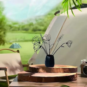 May include: A black metal floral sculpture in a small, dark blue vase. The sculpture features various flower designs. The vase sits on a wooden slab, with a camping scene in the background. A vintage camera is visible on the right.