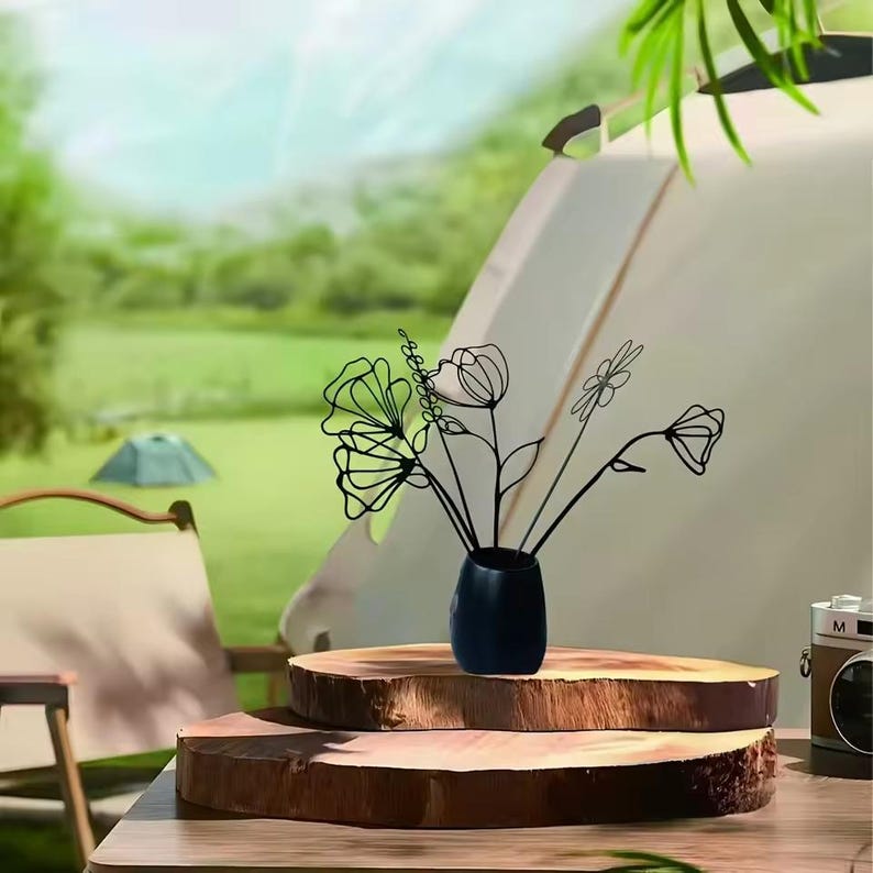 May include: A black metal floral sculpture in a small, dark blue vase. The sculpture features various flower designs. The vase sits on a wooden slab, with a camping scene in the background. A vintage camera is visible on the right.