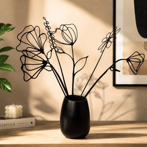 May include: A black ceramic vase holds black metal floral sculptures. The vase has a matte finish and a rounded shape. The metal flowers have a delicate, line-art style, with various flower and leaf designs. A candle and a book are in the background.