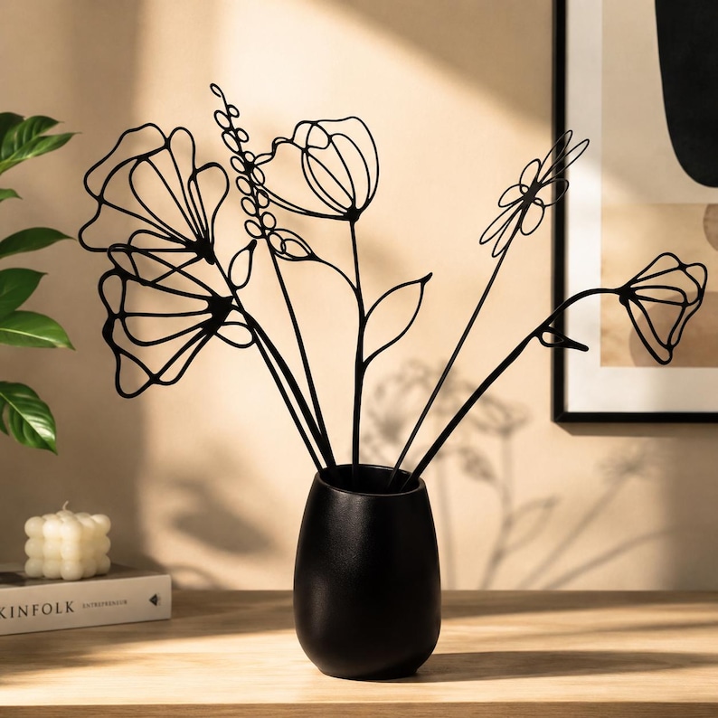 May include: A black ceramic vase holds black metal floral sculptures. The vase has a matte finish and a rounded shape. The metal flowers have a delicate, line-art style, with various flower and leaf designs. A candle and a book are in the background.