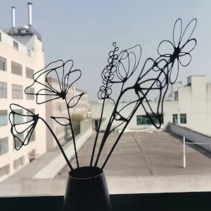 May include: A black metal floral arrangement in a dark vase. The arrangement features several stylized flowers and stems with a minimalist, line-art design. The vase is dark and cylindrical, set against a backdrop of a cityscape.