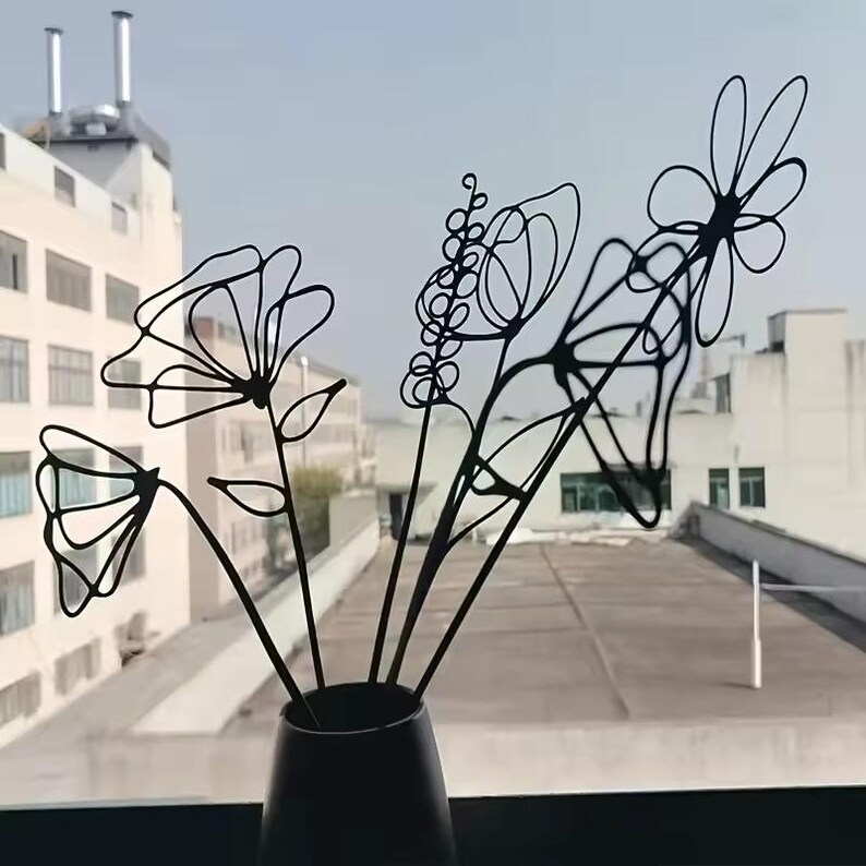 May include: A black metal floral arrangement in a dark vase. The arrangement features several stylized flowers and stems with a minimalist, line-art design. The vase is dark and cylindrical, set against a backdrop of a cityscape.
