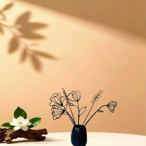 May include: A black vase holds wire flowers with a minimalist design. A white flower with green leaves rests on a piece of driftwood. The background is a warm, neutral color with a leaf shadow.