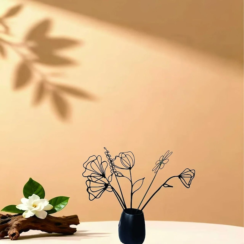 May include: A black vase holds wire flowers with a minimalist design. A white flower with green leaves rests on a piece of driftwood. The background is a warm, neutral color with a leaf shadow.