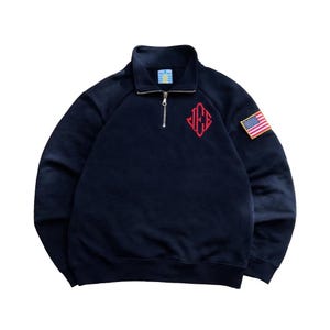 May include: Dark navy blue sweatshirt with a quarter-zip neckline and a red embroidered logo. An American flag patch is on the sleeve. The sweatshirt features a high collar.