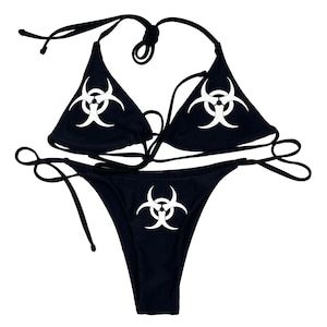 May include: A black bikini set featuring a halter top and a low-rise bottom. Each piece is adorned with a white biohazard symbol. The bikini has black ties for an adjustable fit.