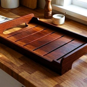 Puede incluir: Bandeja de madera rectangular de color marrón oscuro. Presenta un asa y ranuras paralelas. Se encuentra sobre una encimera de madera, con una toalla a rayas blancas y azules cerca. Se ve una pequeña planta en el fondo.