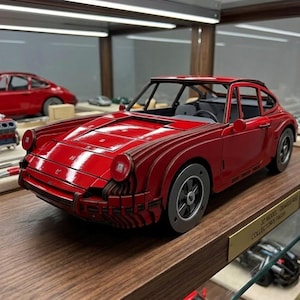 May include: A red model car with a detailed design, displayed on a wooden shelf. The car has a sleek, aerodynamic shape with black wheels and a black interior. Other model cars and car parts are visible in the background.