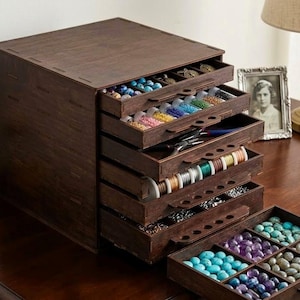 May include: A dark brown wooden storage cabinet with multiple pull-out drawers designed for organising craft supplies. The drawers contain various colourful beads, spools of thread, and metal tools, with one drawer fully extended to show compartments.