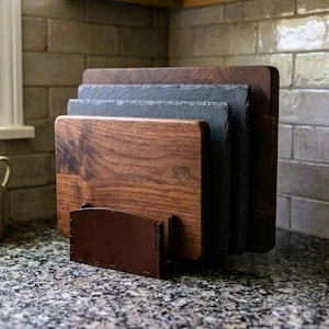 May include: A set of wooden cutting boards in varying sizes and a slate board are displayed in a brown wooden holder. The boards are made of dark wood and slate. The kitchen counter has a granite pattern.