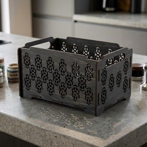 Puede incluir: Una caja de almacenamiento de madera gris oscuro con cortes geométricos. La caja tiene un asa y está sobre una encimera con botes de especias, un prensador de café y una pequeña suculenta en maceta. Almacenamiento de cocina.