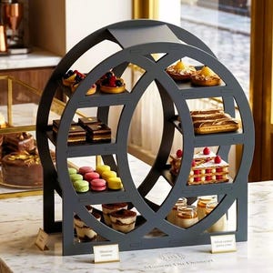 Può includere: Un espositore circolare grigio scuro pieno di vari pasticcini, tra cui crostate, torte e macarons. L'espositore è posto su un piano di lavoro in marmo. Lo sfondo include una macchina da caffè e una finestra.